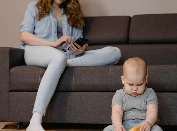 Maman assise sur le canapé du salon, smartphone en main, enregistrant l'historique de vaccination de la famille pendant que ses enfants jouent au sol sur le tapis